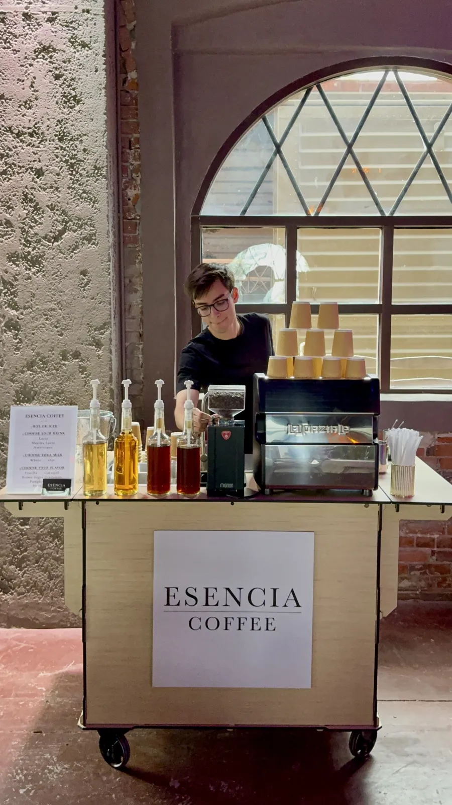 An Esencia Coffee barista working the mobile coffee cart under an arched window at a wedding