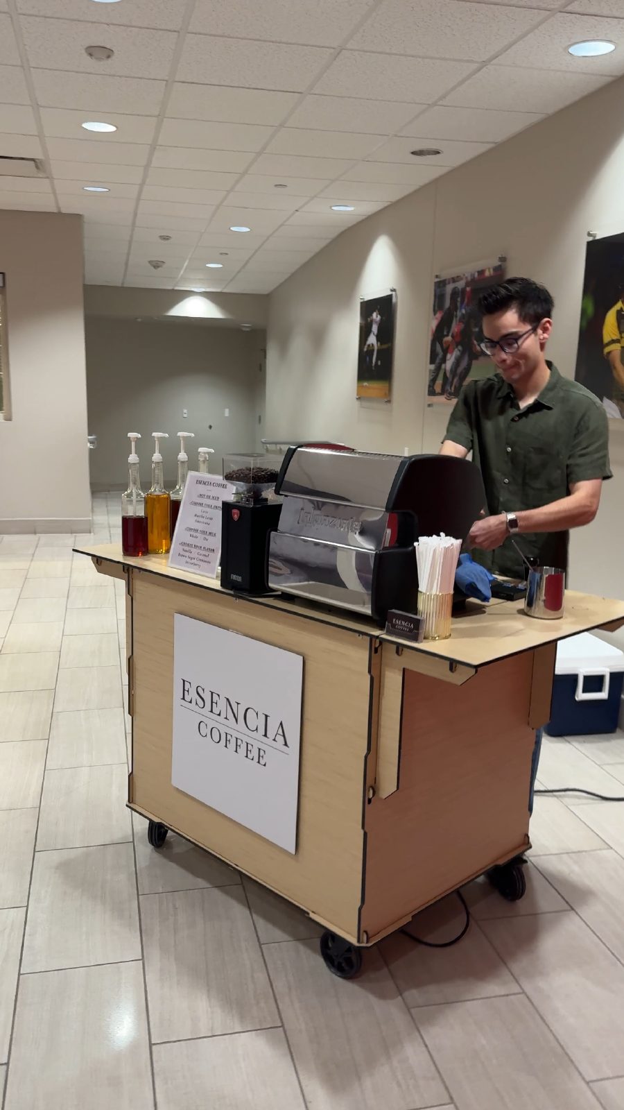 An Esencia Coffee barista making drinks at the cart in a clubhouse at Southwest University Park, with baseball photographs on the wall behind
