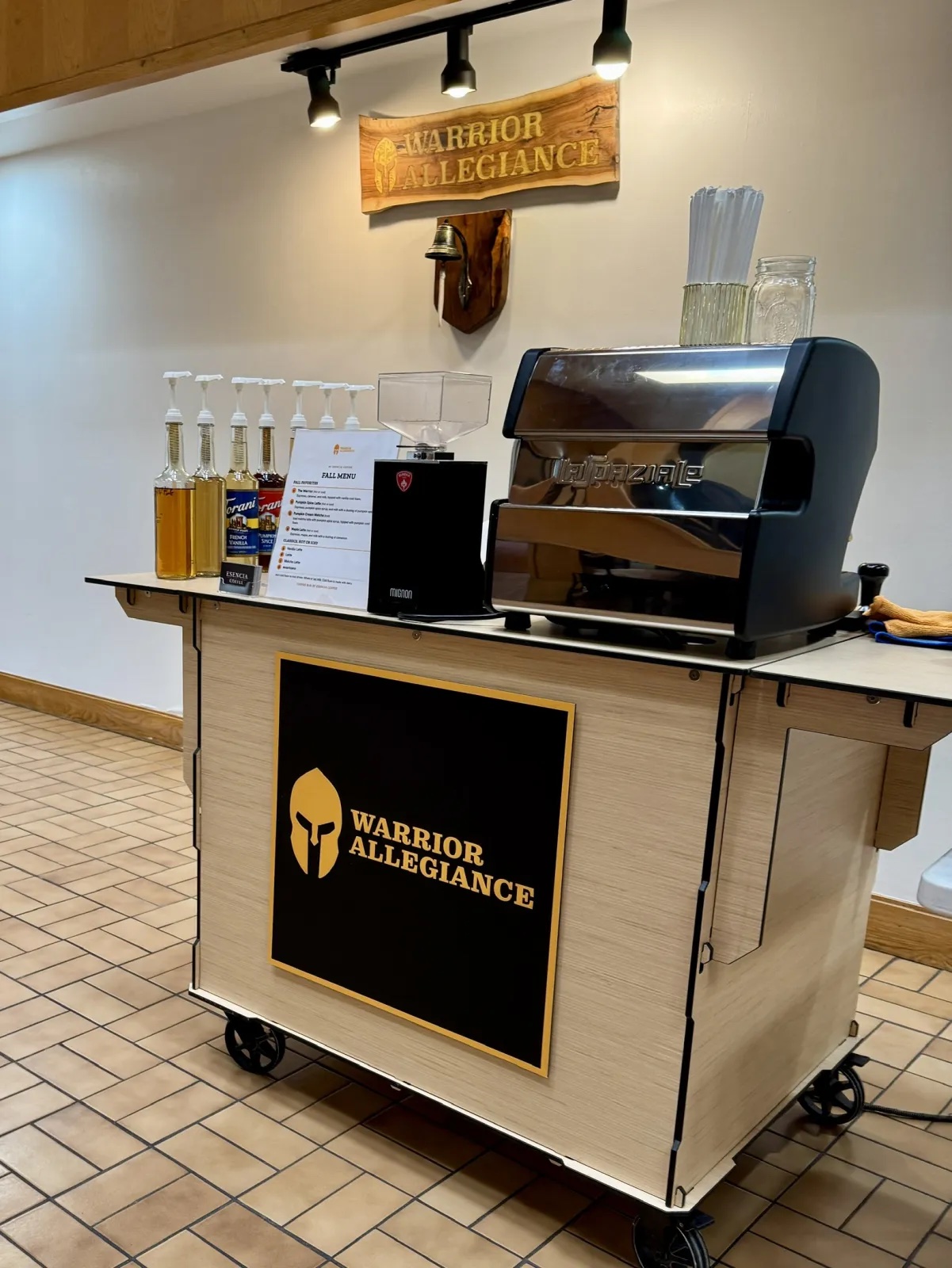 Esencia Coffee cart with the Warrior Allegiance logo on the front and a printed fall menu card beside the espresso machine