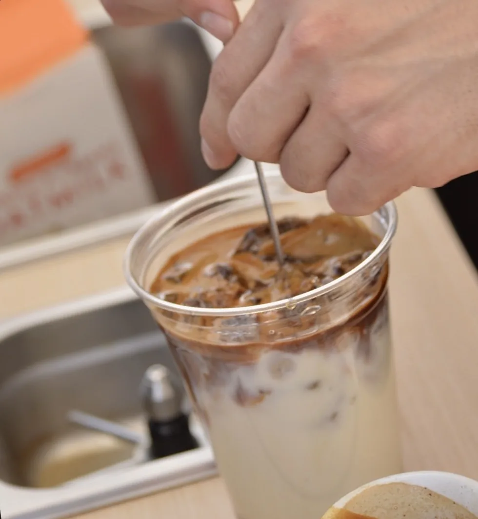 A barista stirring a layered iced latte in a clear cup at an Esencia Coffee event bar.